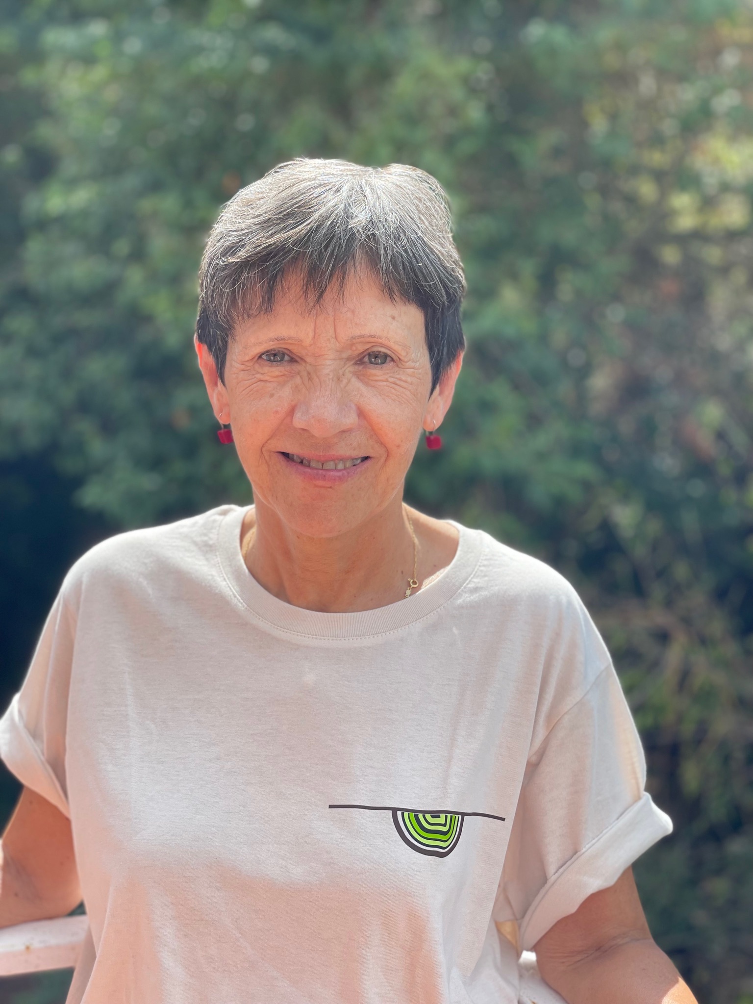 Mujer con cabello corto y canoso sonriente, vestida con una camiseta clara con un diseño colorido en el pecho, de pie frente a un fondo natural difuminado.